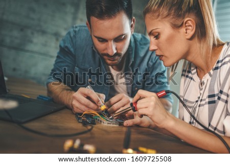 Two young engineers are focused on the repair of electronic equipment by soldering iron. Man is helping woman soldering elements of circuit board. 