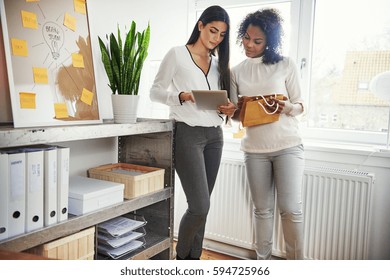 Two Young Entrepreneurs Discussing Packaging Together As They Stand Working On A Design Or Branding On A Tablet For Their Small Business, Shelves Of Binders In The Foreground