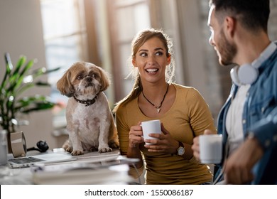 Two young entrepreneurs drinking coffee and communicating in the office while their dog is sitting on a desk. Focus is on smiling woman.