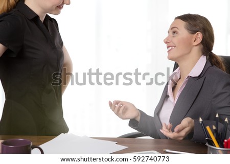 two young, female co-workers in an office.  One sitting at a desk wearing a suit and another standing opposite, having a conversation. 