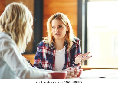Two young female friends chatting over coffee in cafe. Blonde women discussing issues