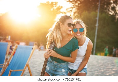 Two Young Female Friends Hangout At The  Beach ,singing And Relaxing In Beautiful Summer Sunset.They Hug Each Other.Best Friends.