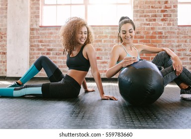 Two Young Female Friends Sitting On Gym Floor With Medicine Ball. Young Woman Working Out In Gym.