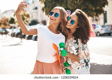 Two young female  hippie brunette and blond women. Models in hipster clothes taking selfie photos for social media on smartphone on the street background. With colorful penny skateboards