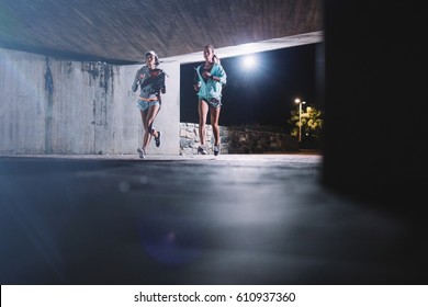 Two Young Female Runners Running Together At Night In City. Young Women Jogging Under Bridge At Night.