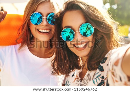 Two young female smiling hippie brunette and blond women models in hipster clothes taking selfie photos for social media on smartphone on the street background. With colorful penny skateboards 