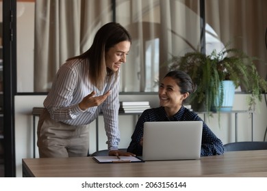 Two young females colleagues of diverse ethnicities discuss job chat hold pleasant conversation at office workplace. Young woman project manager assist indian coworker explain work in corporate app