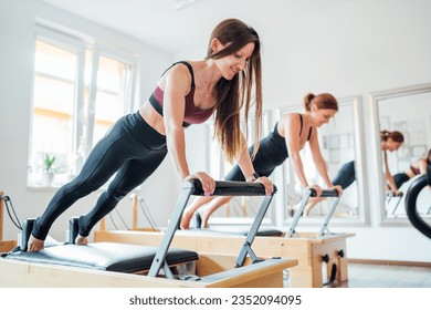 Two young females doing extended plank static strengthening core muscles exercise using pilates reformer machine in sport athletic gym hall. Active people training, yoga classes concept.  - Powered by Shutterstock