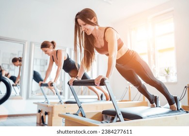 Two young females doing extended plank static strengthening core muscles exercise using pilates reformer machine in sport athletic gym hall. Active people training, yoga classes concept. - Powered by Shutterstock