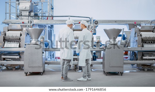Two Young Food\
Factory Employees Discuss Work-Related Matters. Male Technician or\
Quality Manager Uses a Tablet Computer for Work. They Wear White\
Sanitary Hat and Work\
Robes.
