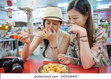 Two Young Foreigner Female Travelers Trying Stinky Tofu With Chopsticks Taiwan Exotic Food Sitting In Street Vendor In Local Market. Girls Covering Nose By Hands With Not Good Smell From Specialty.