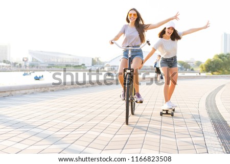 Two young friends relaxing on a hot summer day with one young woman riding a bicycle towing her friend on a skateboard down an urban street.