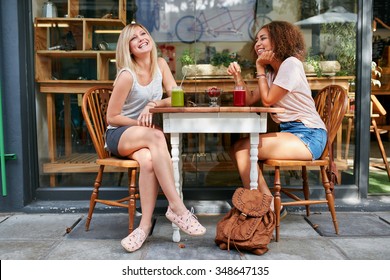 Two Young Friends Sitting At Outdoor Cafe And Smiling. Multiracial Women Hanging Out At Sidewalk Restaurant.