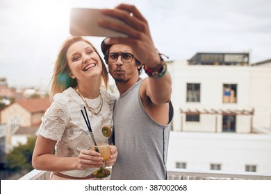 Two young friends taking a selfie on rooftop. Man holding smart phone and taking self portrait with woman holding a cocktail during a party. - Powered by Shutterstock