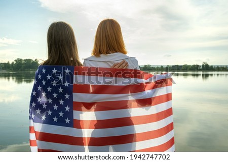 Two young friends women with USA national flag on their shoulders hugging together outdoors on lake shore. Patriotic girls celebrating United States independence day.
