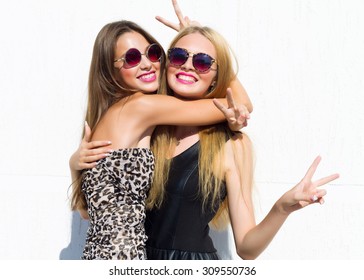 Two young girl friends standing together and having fun,showing signs with their hands. Going crazy,positive emotions.Looking at camera.Two pretty young women in trendy sunglasses and cocktail dresses