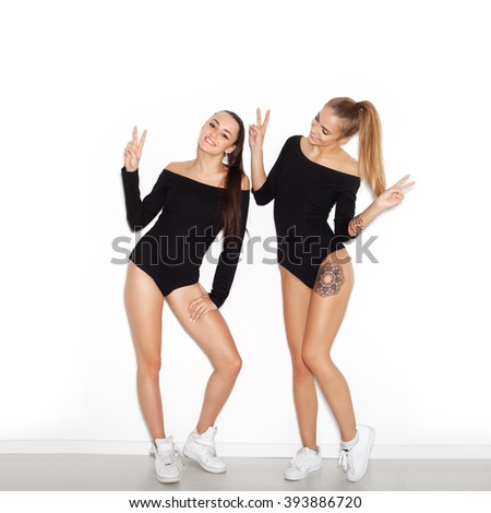 Two young girl friends standing together and having fun. Showing signs with their hands. Looking at camera and smiling. Inside