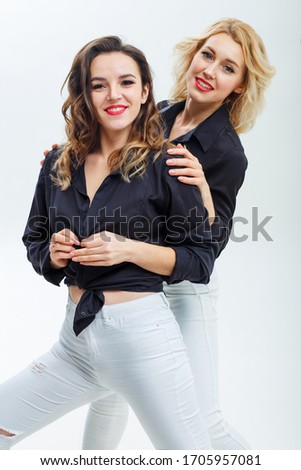 Two young girls in black shirts and white trousers are hugging and smiling at the camera on a white background. Cheerful brunette and blonde