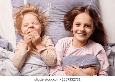 Two young girls lie in bed under soft blankets, sharing joyful moments. The older girl smiles brightly while the younger one covers her mouth, giggling in a cozy bedroom atmosphere. - Powered by Shutterstock