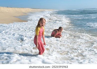 Two young girls are playing in the ocean waves on a sandy beach, laughing and splashing in the foamy water. The sun is shining brightly, creating a fun and carefree summer scene - Powered by Shutterstock