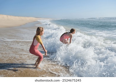 Two young girls are playing in the ocean waves on a sandy beach, laughing and splashing in the foamy water. The sun is shining brightly, creating a fun and carefree summer scene - Powered by Shutterstock