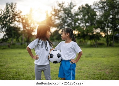 Two young girls are standing in a grassy field, one of them holding a soccer ball. They are both wearing white shirts and blue shorts. Scene is playful and lighthearted - Powered by Shutterstock