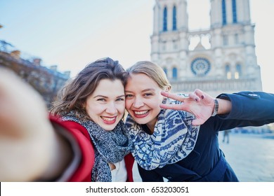 Two young girls walking together in Paris taking selfie with mobile phone near Notre-Dame cathedral. Tourism or friendship concept