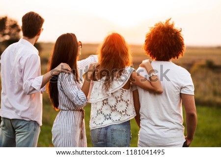 Two young guys and two girls dressed in a stylish clothes are standing in the field and looking in front of them on a sunny day.