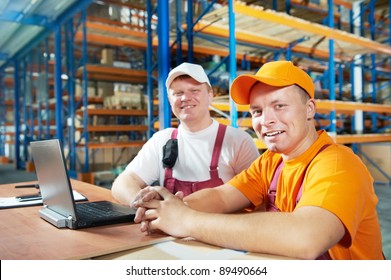 Two Young Handsome Workers Man In Uniform In Front Of Warehouse Rack Arrangement Stillages Using Notebook Computer