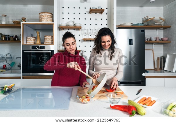 Two\
young happy college student roommates or business women cooking\
food together at their apartment. Females having fun together\
preparing meal in their modern kitchen at cozy\
home.