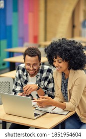 Two Young Happy Diverse Colleagues Looking At Laptop And Discussing Something While Sitting At The Desk In Coworking Space