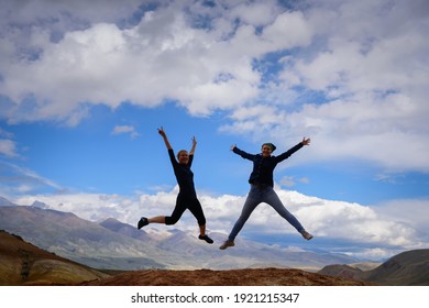 Two young happy girls jump raising hands up with spectacular view of mountains. Female travellers full of joy against the mountains and blue sky with white clouds. Lifestyle in motion concept.