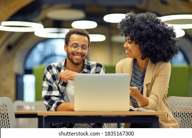 Two Young Happy Multiracial Business People Sitting At The Desk In The Modern Office, Using Laptop And Discussing Project, Man And Woman Working Together In Coworking Space