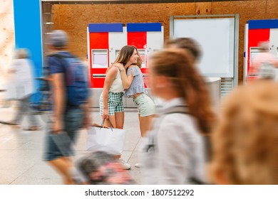 Two young happy smiling women wearing light summer clothes with shopping bags hugging each other meeting on a train station after arrival near many walking people in motion and red ticket machines - Powered by Shutterstock