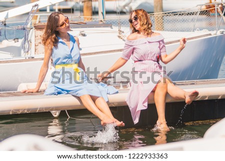 Two young happy smiling women (blonde and brunette) put their feet in the sea - girls are sitting on the pier barefooted and playing with water - rich lifestyle