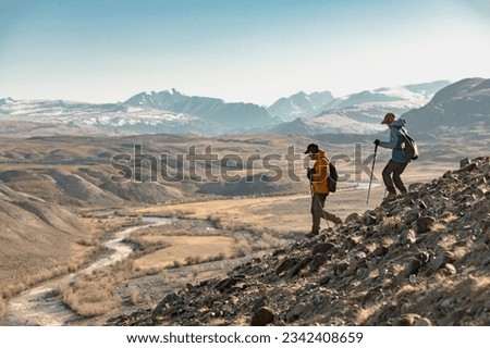 Two young hikers with backpacks and hiking poles walk downhill in mountains. Active tourism in mountains