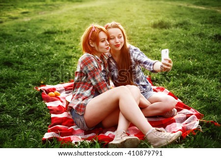 Two Young Hipster Girls Having Fun and Taking Photos (Making Selfie) on Smartphone at the Picnic in a Summer Park at the Sunset. 