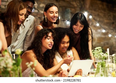 Two Young Intercultural Brides And Their Friends Communicating In Video Chat While Sitting By Served Festive Table During Wedding Feast