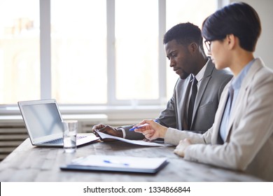 Two Young Intercultural Business Employees In Formalwear Sitting By Desk In Office And Discussing Papers