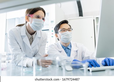 Two young intercultural lab workers in masks, eyeglasses, gloves and whitecoats looking at computer screen during scientific research
