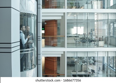 Two young intercultural managers having talk while moving upwards in elevator inside large modern business center with offices