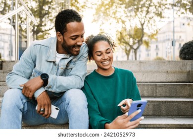 Two young latin american people using smart phone together sitting outdoors. Cheerful african american friends laughing while looking at cellphone screen. Social media concept. Copy space. - Powered by Shutterstock