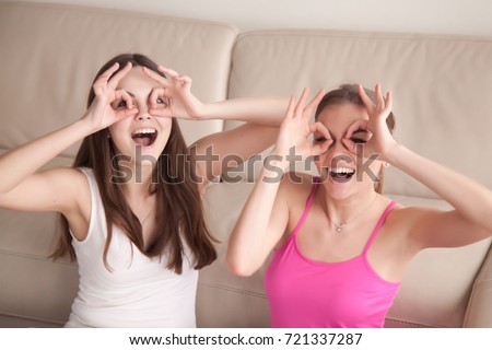Two young laughing girlfriends being silly, constructing goggles with fingers circling their eyes, making funny faces. Best friends having fun and good time at home. Close up, facing camera portrait. 