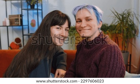 Two young lesbian women family couple looking at camera and smiling at home living room. Loving female gay girls hugging, relaxing together. LGBT people. Romantic love relationship concept and emotion