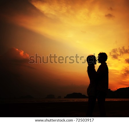 Two young lovers standing on a beach and looking to each other on sunset background