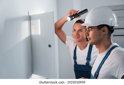 Two young male electricians works indoors together. Using flashlight.