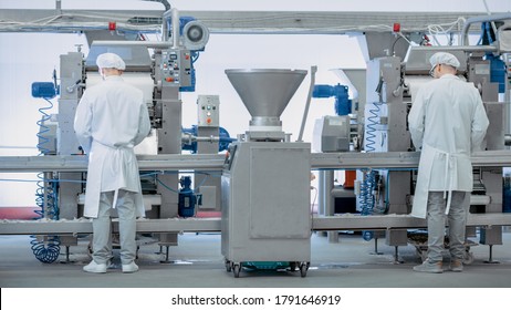 Two Young Male Food Conveyor Belt Employees Work at a Dumpling Factory. They Stand with Their Backs to Camera and Produce Manual Labour on the Line. They Wear White Sanitary Hats and Work Robes.