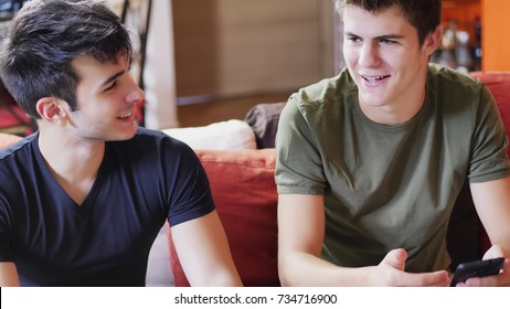 Two Young Male Friends Talking And Chatting While Sitting On Sofa At Home, Relaxing And Having Fun Together.