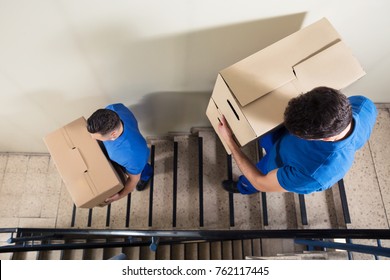 Two Young Male Movers In Blue Uniform Carrying Cardboard Boxes On Staircase