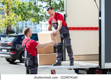 Two Young Male Movers In Uniform Carrying Cardboard Boxes From Truck
